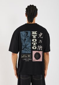 T-shirt noir oversize avec un design graphique comportant des éléments bleus et roses, y compris du texte japonais et des motifs floraux au dos.