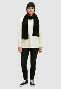 Berretto a costine nero, sciarpa e maglione oversize sopra jeans skinny neri. Sneakers con suola bianca. Dettaglio del logo su berretto e sciarpa.