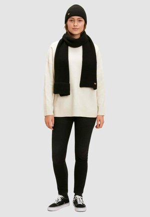 Berretto a costine nero, sciarpa e maglione oversize sopra jeans skinny neri. Sneakers con suola bianca. Dettaglio del logo su berretto e sciarpa.