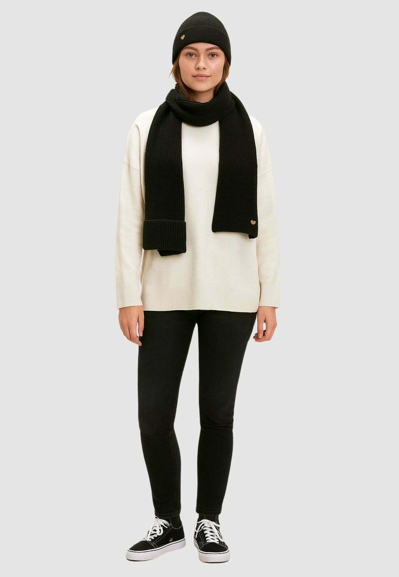 Berretto a costine nero, sciarpa e maglione oversize sopra jeans skinny neri. Sneakers con suola bianca. Dettaglio del logo su berretto e sciarpa.