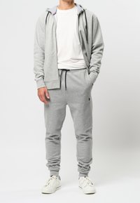 Hellgrauer Hoodie und Jogginghose aus weichem Baumwollmischgewebe mit Fronttasche, Tunnelzug an der Taille und gerippten Bündchen. Weiße Turnschuhe vervollständigen das Outfit.