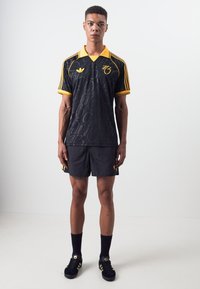 Maillot de sport noir avec des accents jaunes, présentant un design à motifs, associé à un short noir et des chaussures de sport noires avec trois bandes.