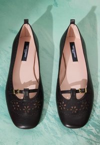 Paire de ballerines Mary Jane en cuir noir avec des perforations en forme de fleur et des brides à boucle dorée sur une surface texturée bleu sarcelle.