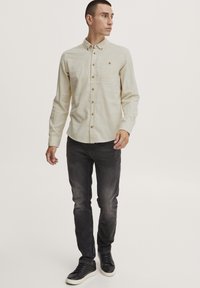 Chemise boutonnée beige clair avec poche poitrine, associée à un jean noir décoloré et des baskets noires. Texture lisse ; aucun motif visible.