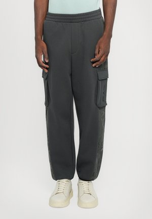 Pantalons cargo gris avec taille élastique, poches latérales et panneaux latéraux texturés. Portés avec des baskets claires.