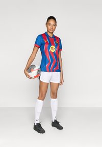 Φανέλα ποδοσφαίρου της FC Barcelona σε μπλε με κόκκινες ζιγκζαγκ ρίγες, λευκό σορτς, μαύρα παπούτσια ποδοσφαίρου και ποδοσφαιρικές επιγονατίδες. Το μοντέλο κρατά μια μπάλα ποδοσφαίρου.