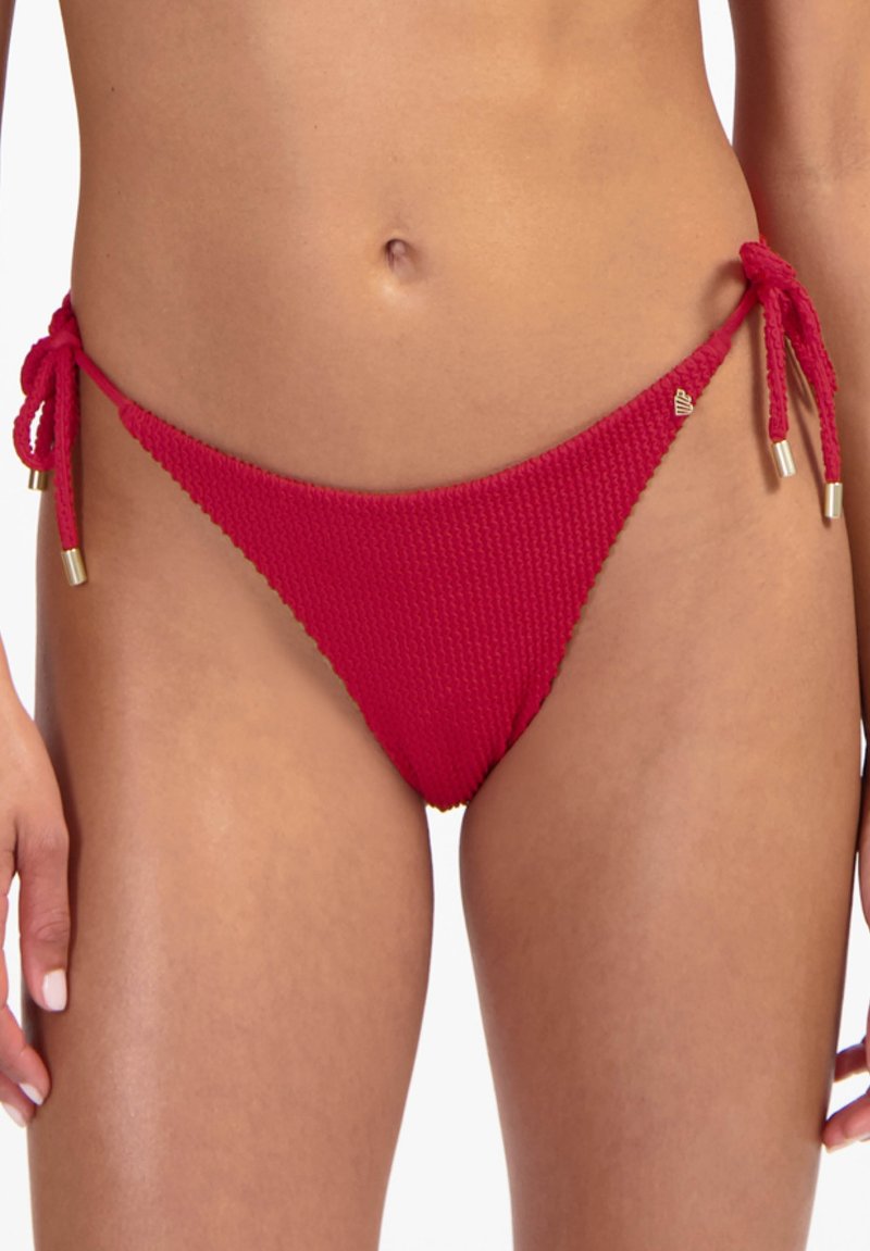Roter Bikinihöschen mit strukturierter Oberfläche, seitlichen Bindungen und Metallakzenten, getragen an den Hüften und dem unteren Bauchbereich einer Person.