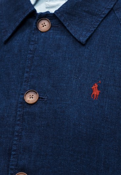 Πουκάμισο ναυτικού μπλε με καφέ κουμπιά και κόκκινο κεντητό λογότυπο παίκτη πόλο στο στήθος, ορατό υφή υφάσματος.