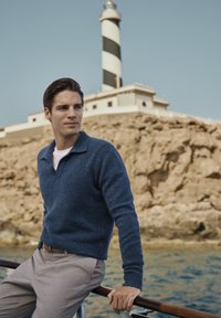 Marineblauwe, gestructureerde trui met een kraag, gecombineerd met lichtgrijze broek. Op de achtergrond een gestreepte vuurtoren op rotsachtig terrein.