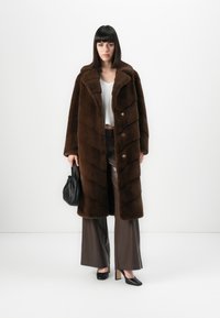 Donna che indossa un lungo cappotto di pelliccia marrone, una maglia bianca, pantaloni larghi marroni, tacchi neri e tiene in mano una piccola borsa nera su uno sfondo bianco.