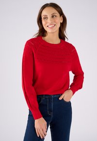 Maglione rosso lavorato a maglia con un giromanica testurizzato, caratterizzato da un design a reticolo sulle maniche. Abbinato a jeans blu, che mostrano un fit rilassato.