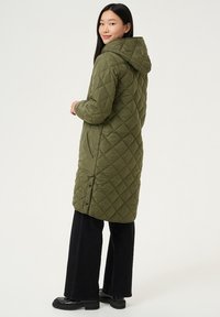 Parka matelassée verte avec capuche, dotée de poches latérales et d'un ourlet légèrement arrondi. Portée avec un pantalon large noir et des bottes épaisses.