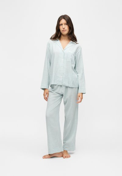 Femme debout pieds nus portant un haut de pyjama à manches longues rayé verticalement bleu clair et blanc avec un pantalon large assorti sur fond blanc.