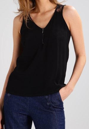 Kobieta w czarnym topie bez rękawów z frontowym zamkiem błyskawicznym i ciemnoniebieskich spodniach na guziki, stojąca z jedną ręką w kieszeni.