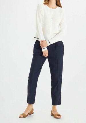 Kvinde iført hvid langærmet sweater med sort kant, marineblå tilspidsede bukser og tan vævede sandaletter med ankelsnor, stående mod hvid baggrund.