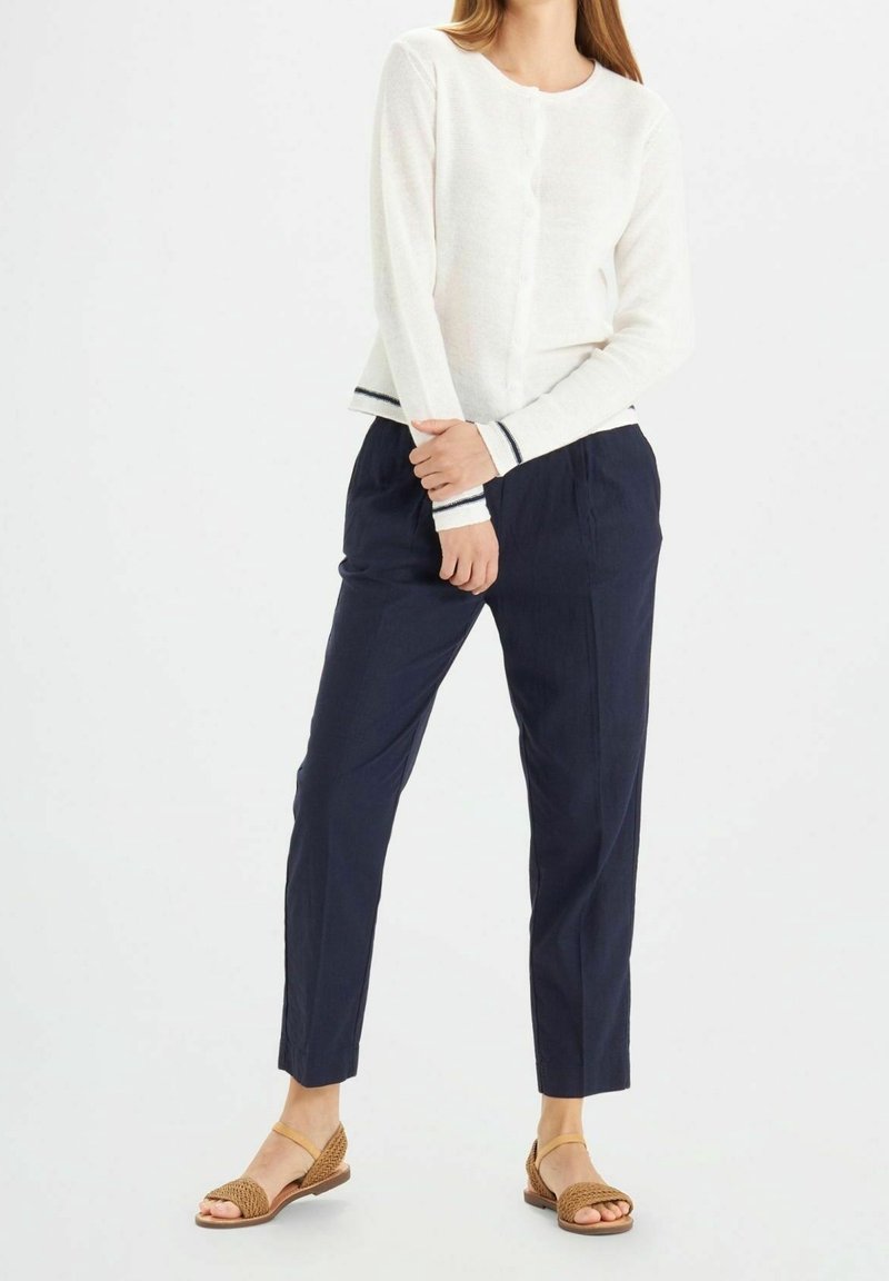 Femme portant un pull blanc à manches longues avec bordure noire, un pantalon bleu marine fuselé et des sandales tressées beige avec une bride autour de la cheville, debout sur un fond blanc.