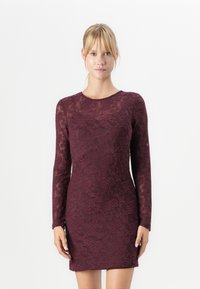 Femme blonde portant une robe en dentelle bordeaux ajustée à manches longues, se tenant devant un fond blanc.