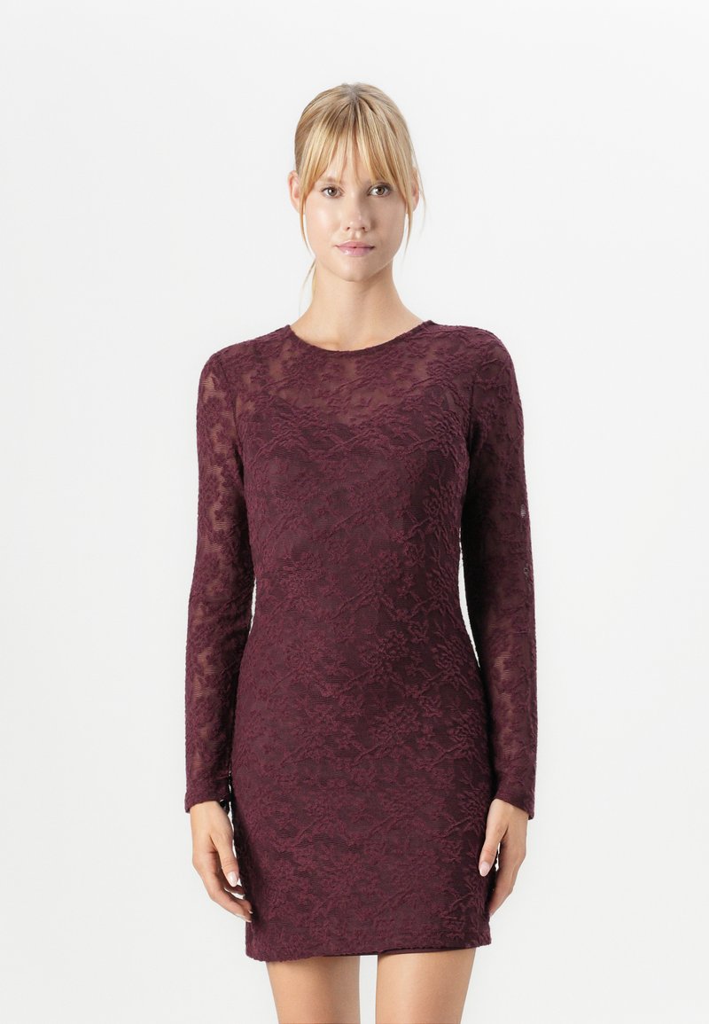 Femme blonde portant une robe en dentelle bordeaux ajustée à manches longues, se tenant devant un fond blanc.