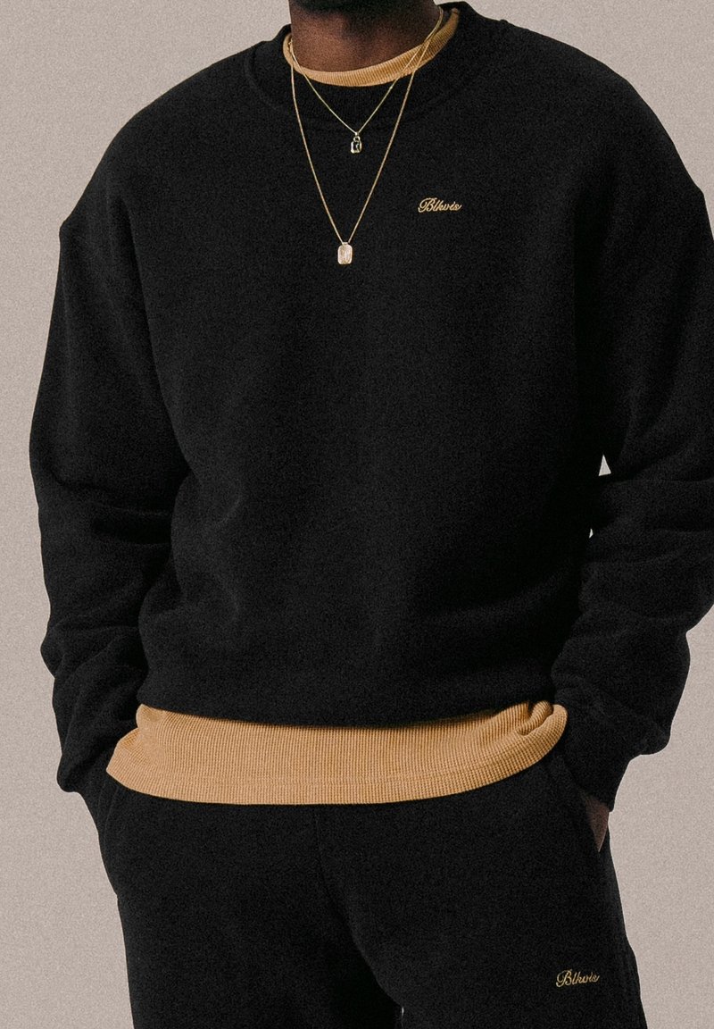 Personne portant un sweat-shirt noir et un pantalon avec un liseré jaune et un logo "Blvker" brodé, accessoirisée de deux colliers en or.