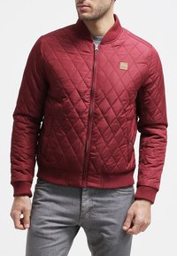 Red quilted bomber jacket with a zip front, ribbed cuffs and hem, featuring a small logo patch on the chest. Smooth fabric texture.