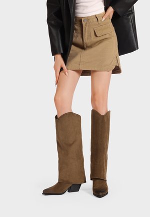 Stivali alti in suede marrone con punte affusolate e bordi angolati, abbinati a una mini gonna in denim khaki con tasche frontali e uno spacco laterale.