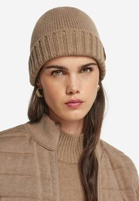 Stickad beige mössa med ribbat mönster. Modellen har på sig en matchande beige tröja med hög krage och en vadderad jacka.