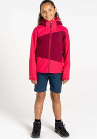 Rote und kastanienbraune wasserdichte Jacke mit Kapuze, ausgestattet mit einem Reißverschluss vorne und elastischen Bündchen, kombiniert mit dunklen Shorts und schwarzen Wanderschuhen.
