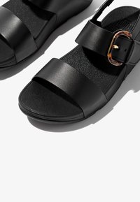 Sandalias negras con dos correas, una brillante y otra mate. Presenta una hebilla de carey. Plantilla negra texturizada con suela plana.