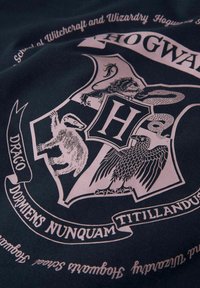 Tissu noir avec un design de blason de Poudlard en rose, affichant un lion, un serpent, un aigle et une blaireau, avec du texte dans une police élégante.