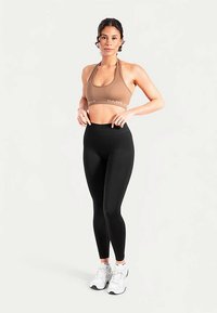 Brassière de sport marron clair avec le texte "SMIL", associée à un legging noir taille haute. Le mannequin porte des baskets blanches et des chaussettes avec des accents de couleur.