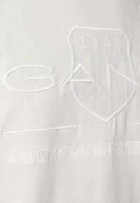 Valkoinen puuvillatee-paita, jossa on kohokuvioitu kilpimalja, jossa on vuosi '1949' ja sanat "GAB American Sports" valkoisella brodeerauksella.