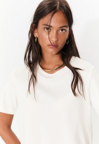 Bijela majica kratkih rukava glatke teksture, s okruglim izrezom i suptilnim detaljima šavova; prikazan višeslojni ogrlica zlatne boje.