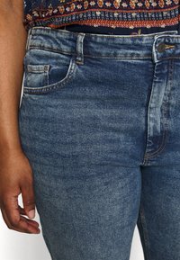 Jeans de mezclilla con un lavado azul oscuro desvanecido, con cintura alta, bolsillo frontal y acentos de costura clara. Textura suave y ajuste clásico.