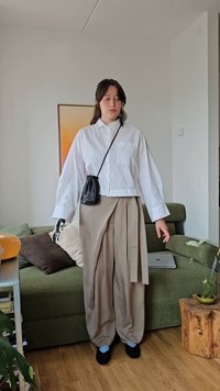 Weißes, gekürztes Hemd mit Knöpfen, beiger, plissierter weit geschnittener Hose, schwarze Umhängetasche und schwarze Schuhe. Der Hintergrund zeigt ein grünes Sofa und eine Pflanze.