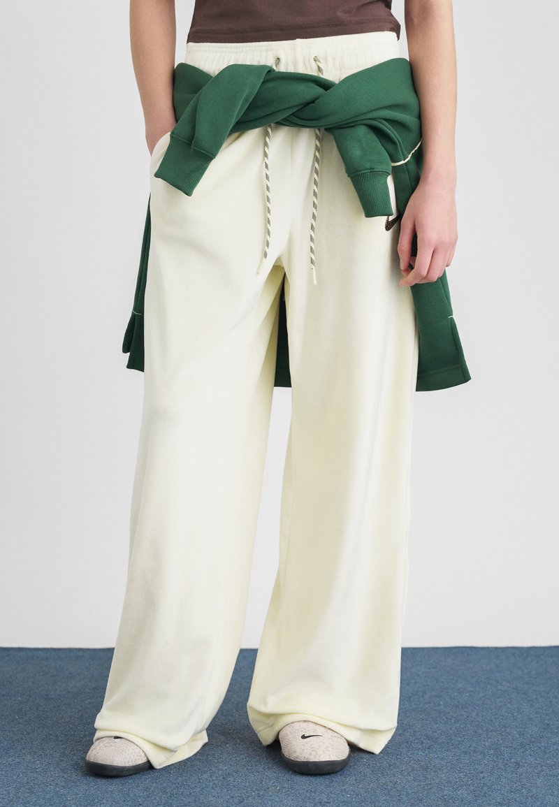 Személy laza krémszínű melegítőnadrágot, szürke Nike félcipőt és egy derékon megkötött zöld pulóvert visel, kék szőnyegen állva.