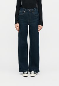 Γυναίκα που φοράει σκούρο μπλε τζιν παντελόνι με ψηλή μέση και φαρδιά μπατζάκια, μαύρο μακρυμάνικο τοπ και ασπρόμαυρα αθλητικά παπούτσια, στέκεται μπροστά σε ένα απλό φόντο.