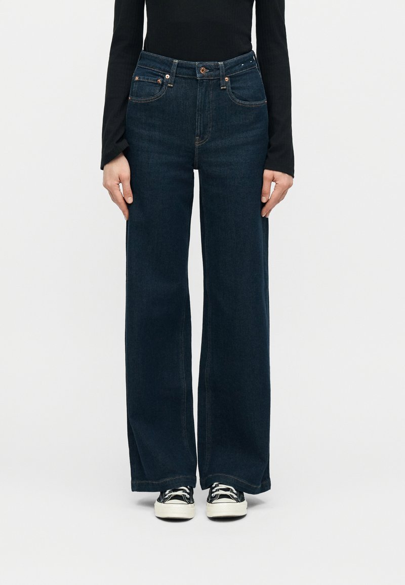 Γυναίκα που φοράει σκούρο μπλε τζιν παντελόνι με ψηλή μέση και φαρδιά μπατζάκια, μαύρο μακρυμάνικο τοπ και ασπρόμαυρα αθλητικά παπούτσια, στέκεται μπροστά σε ένα απλό φόντο.