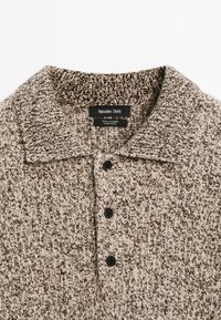 Beige und braun gestricktes Kaschmir-Polohemd mit kragen, fünf schwarzen Knöpfen und einem strukturierten Muster. Etikett am Kragen sichtbar.