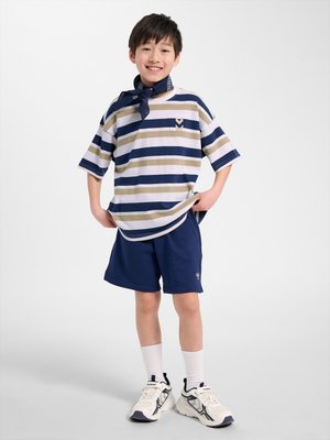 Lachende jongen die marineblauwe shorts, een gestreept beige en marineblauw T-shirt, witte sneakers, witte sokken en een marineblauwe halsdoek draagt, staand met handen in de zij.