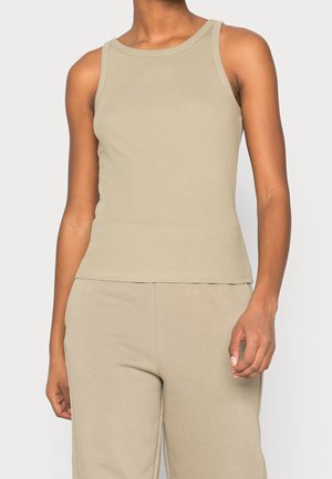 Vrouw draagt een mouwloos beige geribd tanktopje en bijpassende beige broek tegen een effen lichte achtergrond.