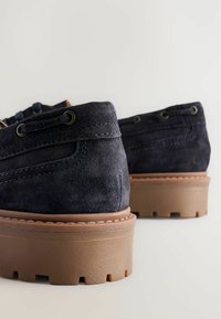 Pantofi din suede albastru închis cu o talpă groasă culoare bej. Au ochiuri metalice și cusături detaliate pe margine.