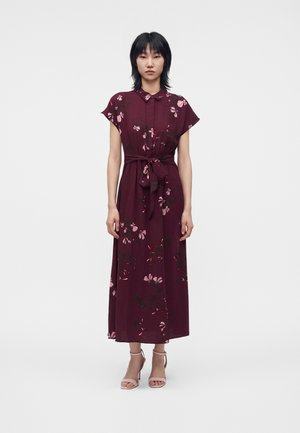 Femme portant une robe sans manches rouge foncé à motif floral avec une ceinture à la taille et des talons ouverts beiges, debout devant un fond clair uni.