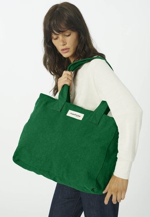 Femme tenant un grand sac cabas vert avec une étiquette blanche "rivedroite", portant un haut à manches longues blanc et un jean bleu foncé sur un fond blanc.