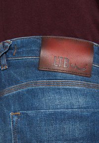 Modré džíny z denimu s hnědým koženým štítkem na pasu s nápisem "LTB." Obsahují viditelné švy a klasický design kapes.