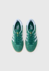 Sapatilhas de camurça verde com detalhes em azul claro, três riscas brancas e sola de borracha cor de goma; marca dourada na língua.