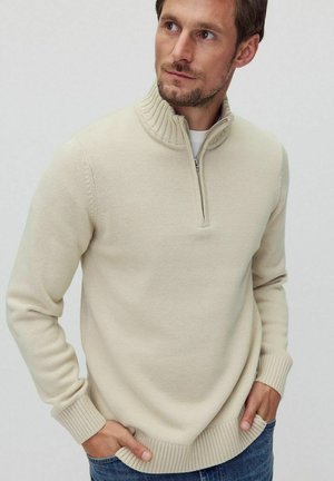 Pull en maille beige à col zippé. Caractéristiques d'un col et de poignets côtelés, avec une coupe décontractée. Associé à un jean bleu foncé.