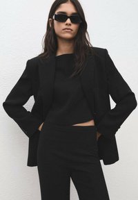 Blazer negro entallado sobre un top negro corto y pantalones ajustados, con líneas limpias y textura suave. La modelo lleva gafas de sol negras.