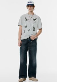 Gry T-shirt med grafiske tryk, løse mørkeblå jeans og beige sko. En afslappet kasket med blå skrift fuldender outfittet.