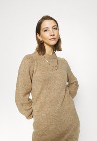 Bež pleten sweatshirt z dolgimi rokavi in rebrastim ovratnikom, ki se ponaša z mehko teksturo. V kombinaciji z večplastnimi zlatimi verižicami.