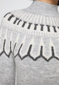 Suéter de punto gris claro con un cuello alto, que presenta un diseño de patrón en blanco y negro en el canesú y una textura de tela.